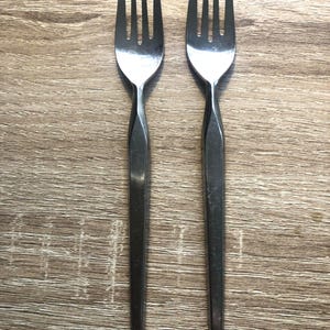 Puede incluir: Dos tenedores de acero inoxidable, cada uno con cuatro púas, dispuestos verticalmente sobre una superficie de madera. Los tenedores tienen un diseño simple y estilizado con mangos largos y delgados.