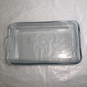 Puede incluir: Una fuente rectangular de vidrio transparente para horno con asas laterales integradas y un patrón decorativo en relieve en la base. El diseño incluye un motivo circular central con volutas intrincadas y bordes lineales en los lados.