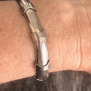 May include: A silver-toned bracelet with a tubular design. The bracelet features a series of connected, curved segments, creating a unique, articulated look. The metal has a polished finish, reflecting light. The bracelet is worn on a wrist.