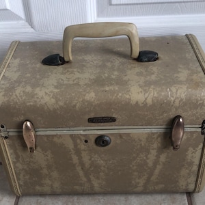 May include: Vintage tan Samsonite train case with a cream-colored handle and brass-toned hardware. The case has a textured finish and a small lock. The Samsonite logo is visible on the front.