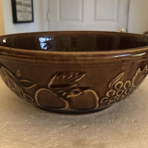 Puede incluir: Un cuenco de cerámica marrón con diseños en relieve de frutas y hojas. El cuenco tiene un borde ancho y un acabado brillante. El diseño de frutas incluye uvas, manzanas y hojas. Este cuenco es adecuado para servir o exhibir.