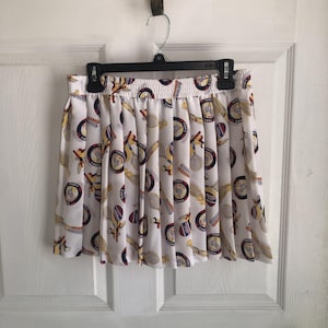 Peut inclure: Une jupe plissée blanche avec un motif imprimé de raquettes de tennis, de balles de tennis et d'emblèmes circulaires portant l'inscription Tennis Sport. La jupe possède une taille élastique et est présentée sur un cintre noir contre une porte.