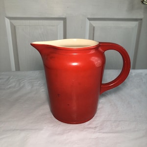 May include: A bright red ceramic pitcher with a smooth, glossy finish. The vessel features a wide, curved pouring spout and a sturdy, integrated handle on the side. The interior of the pitcher is a contrasting cream color, visible at the rim.