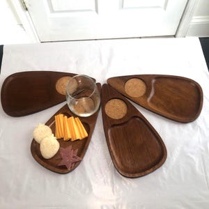 Op de afbeelding: Vier donkerbruine houten serveerplateaus in druppelvorm, elk voorzien van een ronde kurken inleg. Eén plateau bevat een helder glazen beker, terwijl een ander schijfjes gele kaas, twee ronde crackers en drie stervormige stukjes vlees bevat.