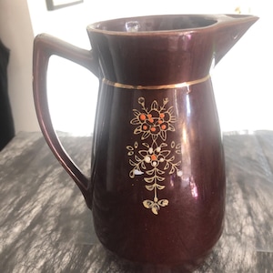 May include: A brown ceramic pitcher with a handle and spout. The pitcher is decorated with gold trim and floral designs in orange and white. The pitcher is likely used for serving beverages.