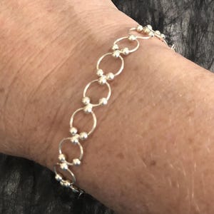 May include: A silver-toned bracelet composed of a series of interconnected circular links. Each circular link is joined to the next by small, spherical silver beads, creating a repeating pattern that wraps around the wrist in a delicate chain design.