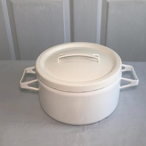 Puede incluir: Una olla redonda de metal esmaltado blanco con tapa a juego. La olla cuenta con dos asas laterales angulares y un asa central en la tapa. El diseño es sencillo y funcional, con un acabado blanco liso y brillante que cubre todo el exterior.