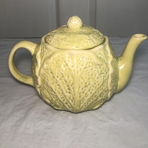 May include: A pale yellow ceramic teapot featuring a textured surface that mimics the appearance of overlapping cabbage leaves. The teapot includes a matching lid with a small knob handle, a curved spout, and a rounded handle on the side.