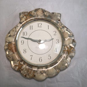 May include: A wall clock shaped like a scallop shell with a clear resin frame embedded with various seashells and starfish. The clock face is white with black Arabic numerals and silver-toned hands. The word "FIRST" is printed on the clock face.