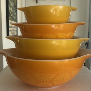Peut inclure: Ensemble de quatre bols à mélanger Pyrex vintage, jaunes et oranges. Le plus grand bol présente un motif de marguerites. Chaque bol a une poignée de chaque côté. Les bols sont empilés les uns dans les autres.