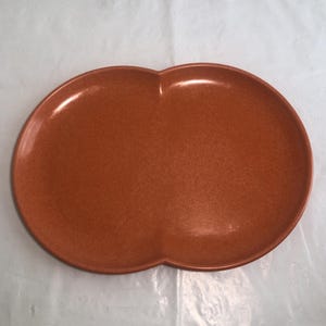 May include: A smooth, terracotta-colored serving platter with a distinct figure-eight or peanut-like shape. The surface features a subtle, speckled texture throughout the entire piece, with a raised rim that follows the curved, indented outline.