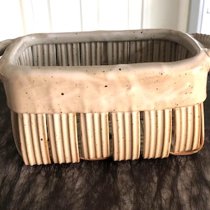 May include: A cream-glazed ceramic basket with a rectangular shape and vertical ribbed detailing. It features two rope-style handles and vertical openings along the sides. The speckled finish gives a rustic appearance to this decorative storage basket.