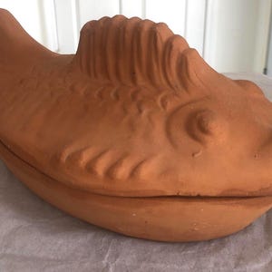 Könnte beinhalten: Eine fischförmige Backform aus Terrakotta mit Deckel. Die Form hat eine warme orangebraune Farbe und weist detaillierte Schuppen, Flossen und eine markante Rückenflosse auf. Der Deckel passt gut.