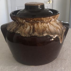 May include: A dark brown ceramic crock with a matching lid and two side handles. The upper portion of the vessel features a decorative drip glaze effect in cream and light brown, creating a textured appearance against the smooth, glossy base.