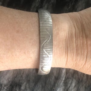 May include: A silver-toned bracelet with a textured surface and a wavy engraved design. The bracelet is flexible and wraps around the wrist. The jewelry piece is a simple, elegant accessory.