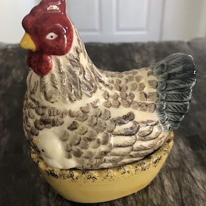 May include: A ceramic chicken figurine sitting in a yellow bowl. The chicken has a red comb, a yellow beak, and white feathers with grey detailing. A small white chick is visible near the base of the chicken. The tail feathers are dark grey.