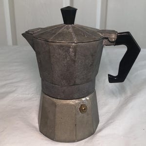 May include: An octagonal stovetop espresso maker constructed from brushed aluminium with a black plastic handle and a matching black knob on the lid. The base features a small brass-coloured pressure release valve on the side of the metal body.