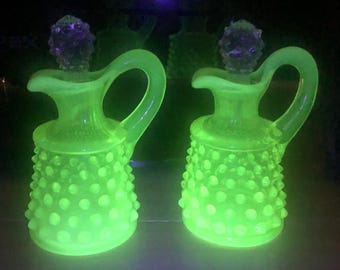 May include: Two matching glass cruets with a vibrant neon green glow under ultraviolet light. Each vessel features a textured hobnail pattern on the base, a curved handle, a narrow neck, and a clear, faceted glass stopper on top.