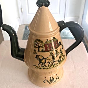 May include: A beige ceramic teapot with a black handle, spout, and top. The teapot features a painted rural scene with a house, trees, and a horse-drawn plow. The base has a decorative green and black design.