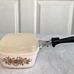May include: A square white ceramic casserole dish with a matching lid, featuring a colorful botanical print of vegetables and herbs on the side. The dish includes a detachable black handle with a chrome-finished metal attachment mechanism.