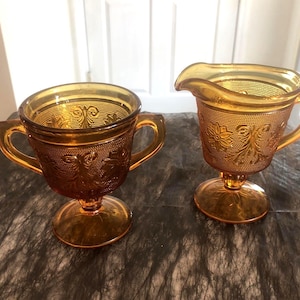 Op de afbeelding: Twee amberkleurige glazen serveerstukken. Eén is een kopje met een handvat, de andere een roomkannetje met een tuit. Beide hebben sierlijke bloemen- en textuurpatronen. De beker heeft twee handvatten en een voet. De roomkannetje heeft één handvat en een voet.