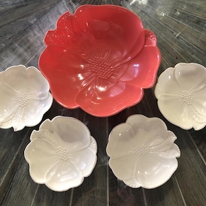 Pode incluir: Um conjunto de travessas de cerâmica com um design floral. O conjunto inclui uma tigela grande, cor de coral, e quatro pratos brancos menores. Cada peça tem a forma de uma flor com detalhes de pétalas e centro.