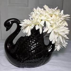 Peut inclure: Un vase ou pot de fleurs en céramique noire brillante en forme de cygne, orné d'un motif de plumes texturé sur le corps. Le cygne contient un bouquet de fleurs artificielles blanches aux pétales fins, formant un ensemble décoratif contrasté.