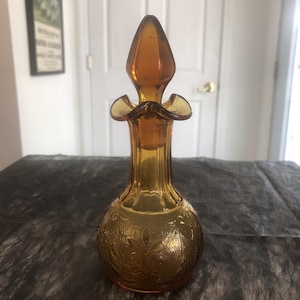 May include: Amber-colored glass perfume bottle with a decorative stopper. The bottle has a bulbous base with an embossed floral pattern and a slender neck. The stopper is pointed and has a flared, petal-like design. The bottle is likely vintage.