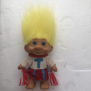 May include: A troll doll with bright yellow hair, tan skin, and blue eyes. The doll wears a white shirt with a blue letter T on the chest, a red and blue skirt, and holds two red and white pom-poms in its hands. The figure has a smiling expression.