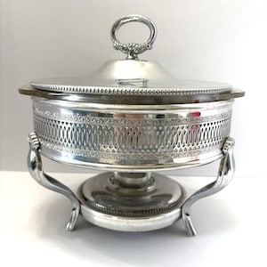 May include: A silver-colored serving dish with a lid and a decorative handle. The dish has a detailed, openwork design around the sides and sits on a matching stand with curved legs. The lid has a decorative rim.