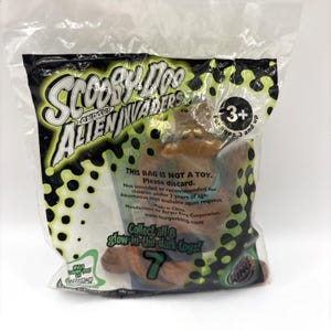 May include: A sealed plastic bag featuring a Scooby-Doo and the Alien Invaders toy. The bag has a green and black design with the text "Scooby-Doo and the Alien Invaders." The toy is a brown figure. The bag also has the text "Collect all 8 glow-in-the-dark toys!"
