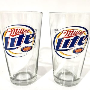 Könnte beinhalten: Zwei transparente Biergläser mit dem Miller Lite-Logo. Das Logo zeigt die Worte "Miller Lite" in Rot und Blau, mit einem goldenen und blauen Oval darum. Die Worte "True Pilsner Beer" stehen am unteren Rand des Logos.