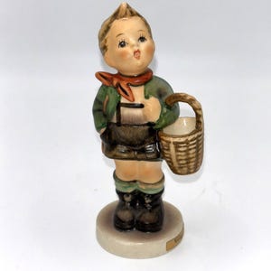 Puede incluir: Figura de cerámica de una persona joven con vestimenta tradicional alemana, sosteniendo una cesta tejida. La figura lleva una chaqueta verde, pantalones cortos marrones, botas negras y una bufanda roja. La figura está sobre una base redonda.