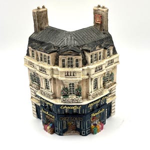 May include: A detailed miniature building with a dark grey roof, beige walls, and multiple windows. The building has a dark blue storefront with the words "Chocolat" and "Chez Louis" visible. Colourful boxes are displayed at the base.