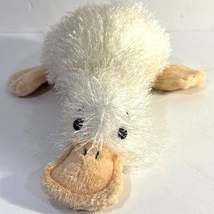 Puede incluir: Un juguete de peluche con forma de ornitorrinco que presenta un cuerpo blanco de textura peluda, con un pico y patas palmeadas de color melocotón claro. El peluche tiene dos ojos pequeños, redondos y negros en la parte frontal de la cabeza.