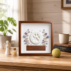 Puede incluir: Un marco cuadrado de madera que contiene una impresión circular de huella de pata en arcilla blanca, flanqueada por flores secas azules y moradas. Una placa de madera oscura bajo la huella tiene grabado el nombre Maverick, 2017-2025 y texto.