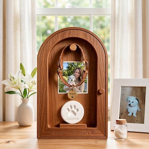 May include: A wooden arched memorial display case featuring a brown leather pet collar, a square photograph of a person with a dog, a round gold tag engraved with the name Cooper and a paw print, and a white clay paw print disc labelled Cooper.