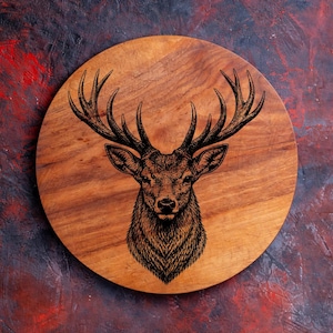 May include: Round wooden cutting board with a detailed engraved image of a stag's head. The board is made of light brown wood and has a dark brown stag illustration in the center. The background is a mix of red, brown, and purple.
