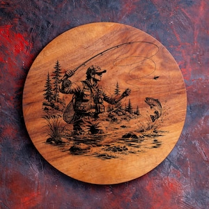 May include: A round wooden board featuring a dark laser-engraved illustration of a person fly fishing in a river. The scene depicts a fisher wearing a vest and hat, casting a line with a rod, while a fish jumps out of the water nearby.