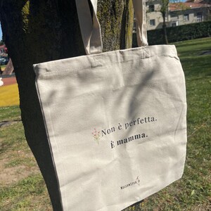 Può includere: Una borsa tote in tela beige con lunghe cinghie, appesa a un albero. La borsa presenta il testo "Non è perfetta. È mamma." in nero, con un piccolo disegno floreale. Il marchio "MELLONSTUDIO" è stampato in basso.
