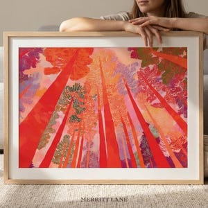 May include: A framed art print featuring a vibrant, colorful illustration of tall trees reaching towards the sky. The artwork showcases a perspective from the forest floor, with red, orange, and pink hues dominating the scene. The text "MERRITT LANE" is at the bottom.