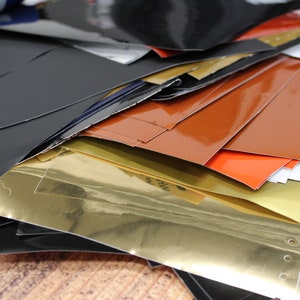 May include: A stack of various coloured vinyl sheets, including black, gold, orange, red, and green. The sheets are glossy and have a smooth surface.