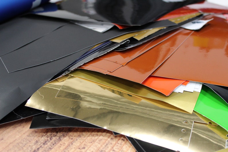 May include: A stack of various coloured vinyl sheets, including black, gold, orange, red, and green. The sheets are glossy and have a smooth surface.