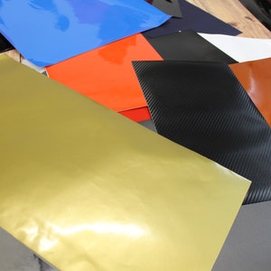 May include: Assortment of colourful vinyl sheets in various colours, including gold, orange, blue, black, and grey. The sheets are arranged in an overlapping pattern, showcasing their different textures and finishes.