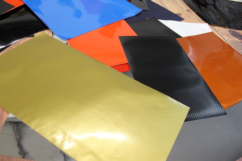 May include: Assortment of colourful vinyl sheets in various colours, including gold, orange, blue, black, and grey. The sheets are arranged in an overlapping pattern, showcasing their different textures and finishes.