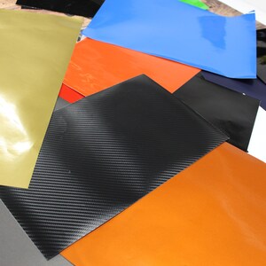 May include: Assortment of vinyl sheets in various colours and textures, including gold, orange, black, blue, grey, and white. The sheets are arranged in a random pattern, showcasing their different finishes.