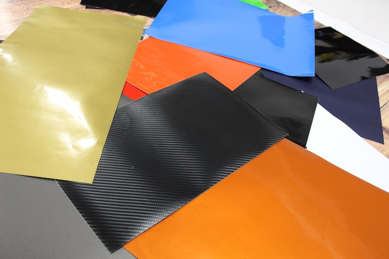 May include: Assortment of vinyl sheets in various colours and textures, including gold, orange, black, blue, grey, and white. The sheets are arranged in a random pattern, showcasing their different finishes.