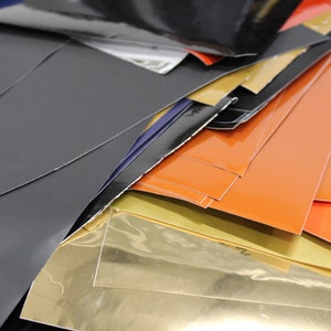 May include: A stack of various coloured vinyl sheets, including black, orange, gold, and grey. The sheets are arranged in a random order, with some overlapping.