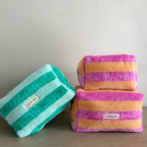 May include: Three colorful, striped rectangular beach towels. One is teal and white, and the other two are pink and orange. Each towel has a small white tag with the text "NOA DE COCO".