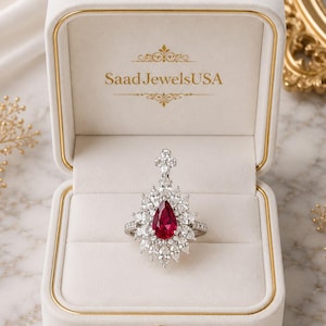 May include: A silver-toned ring featuring a central pear-shaped red gemstone surrounded by a cluster of marquise and round white stones, presented in a cream-colored velvet box with the text SaadJewelsUSA printed in gold on the inner lid.